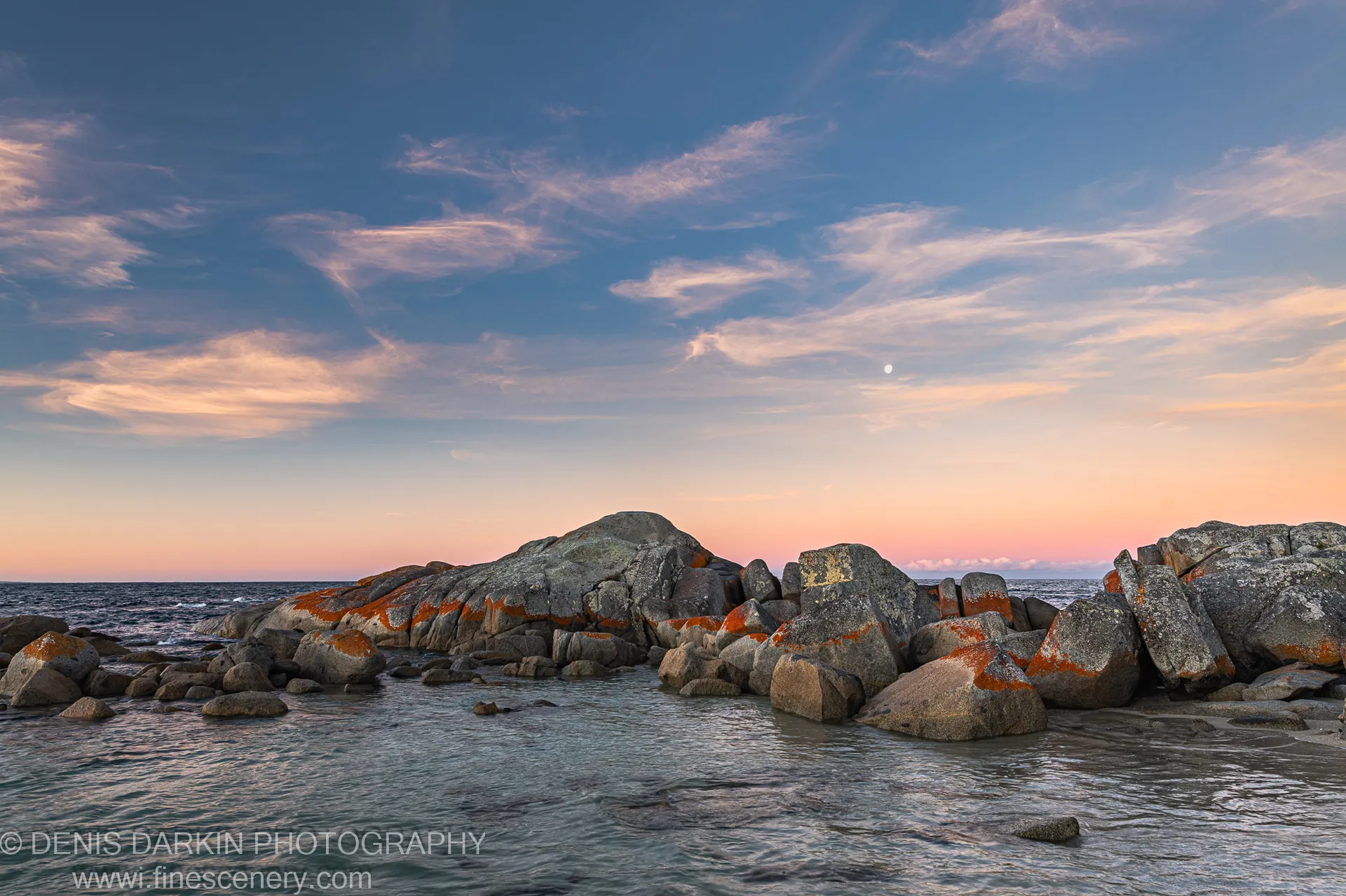 Orange lichen boulders at the tidal edge of Bay of Fires, with a crescent moon visible in the peach evening sky above the water.