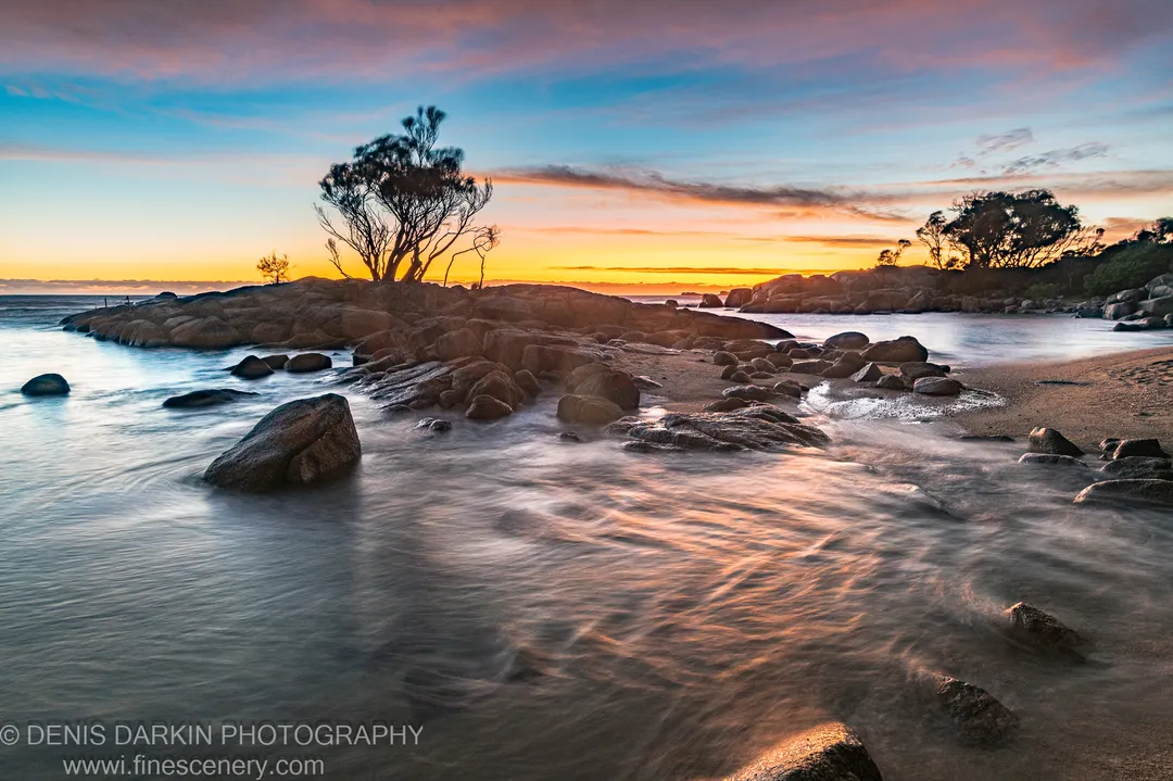 Long-exposure waves move around granite boulders on a sandy Bay of Fires beach, trees silhouetted on the headland in morning light.