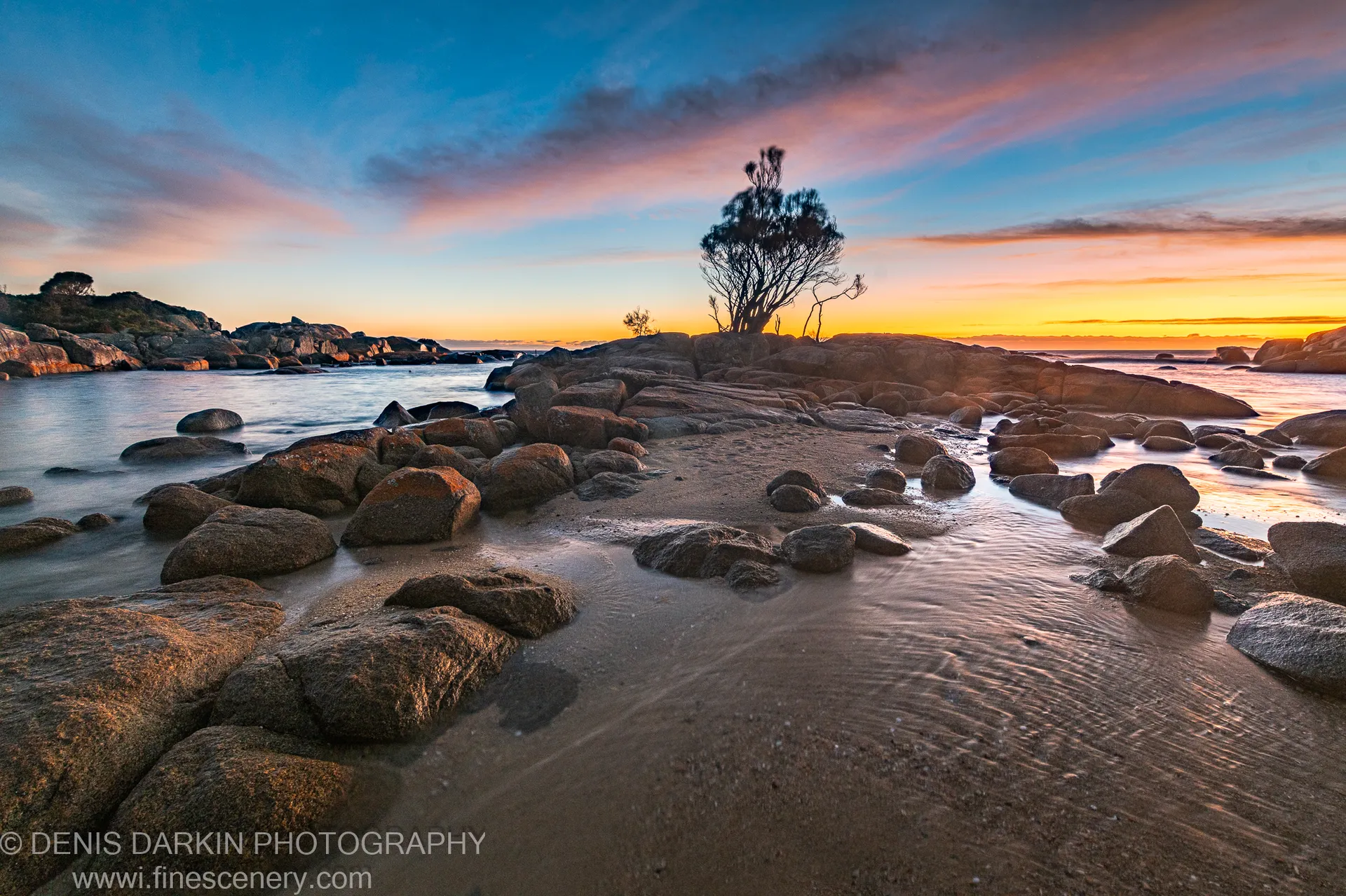 Long-exposure water around granite boulders at Bay of Fires, with a lone tree on a central rock formation and a pink morning sky.