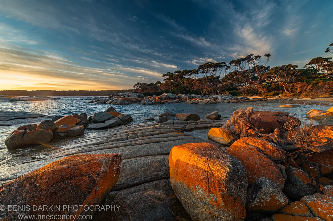 Stratified granite shelves edge a calm body of water leading to a tree-lined headland at Bay of Fires under streaked clouds.
