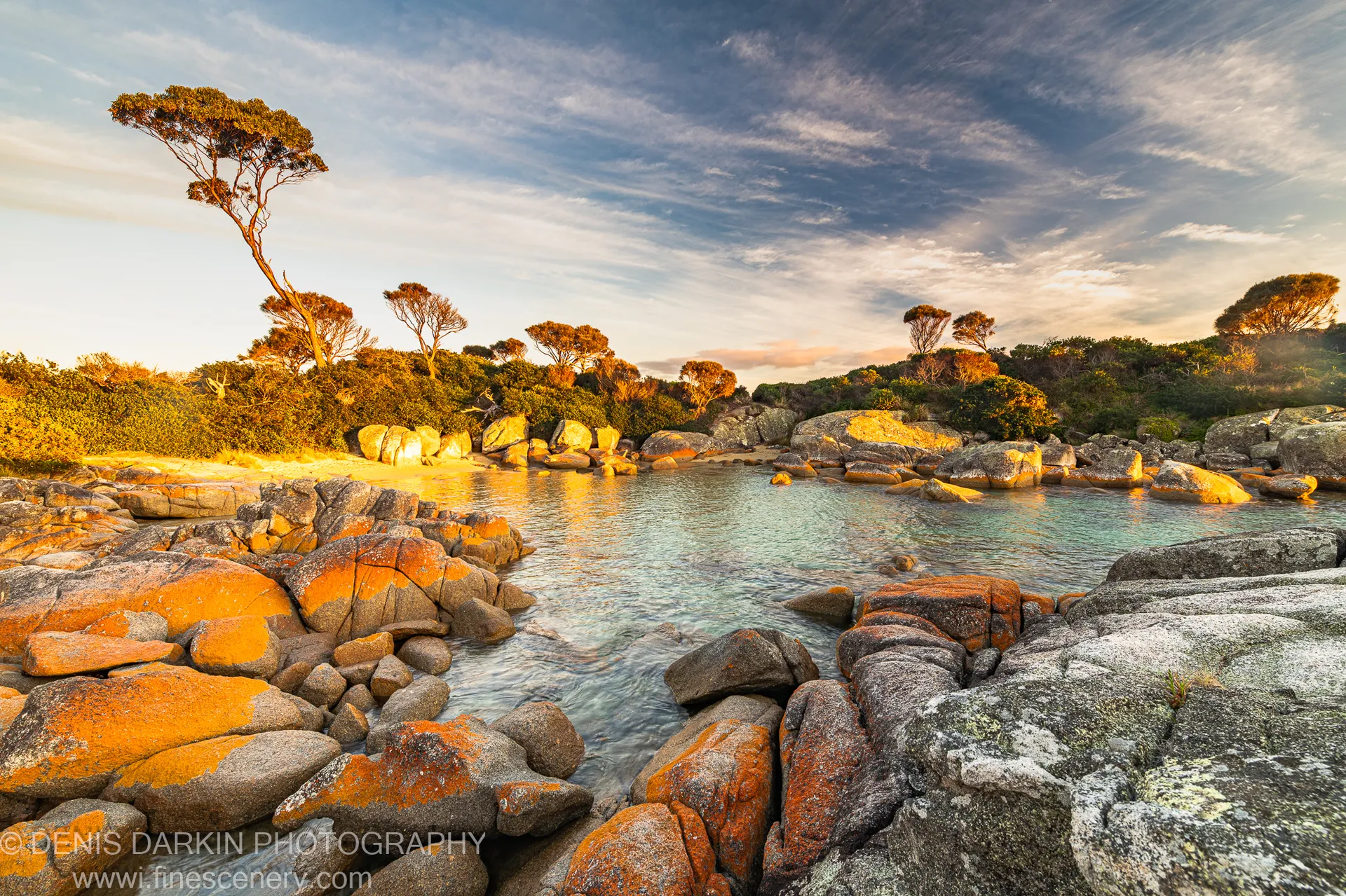 Lichen-covered boulders and golden autumn foliage border a turquoise coastal inlet at Bay of Fires, East Coast Tasmania, under wispy cloud.
