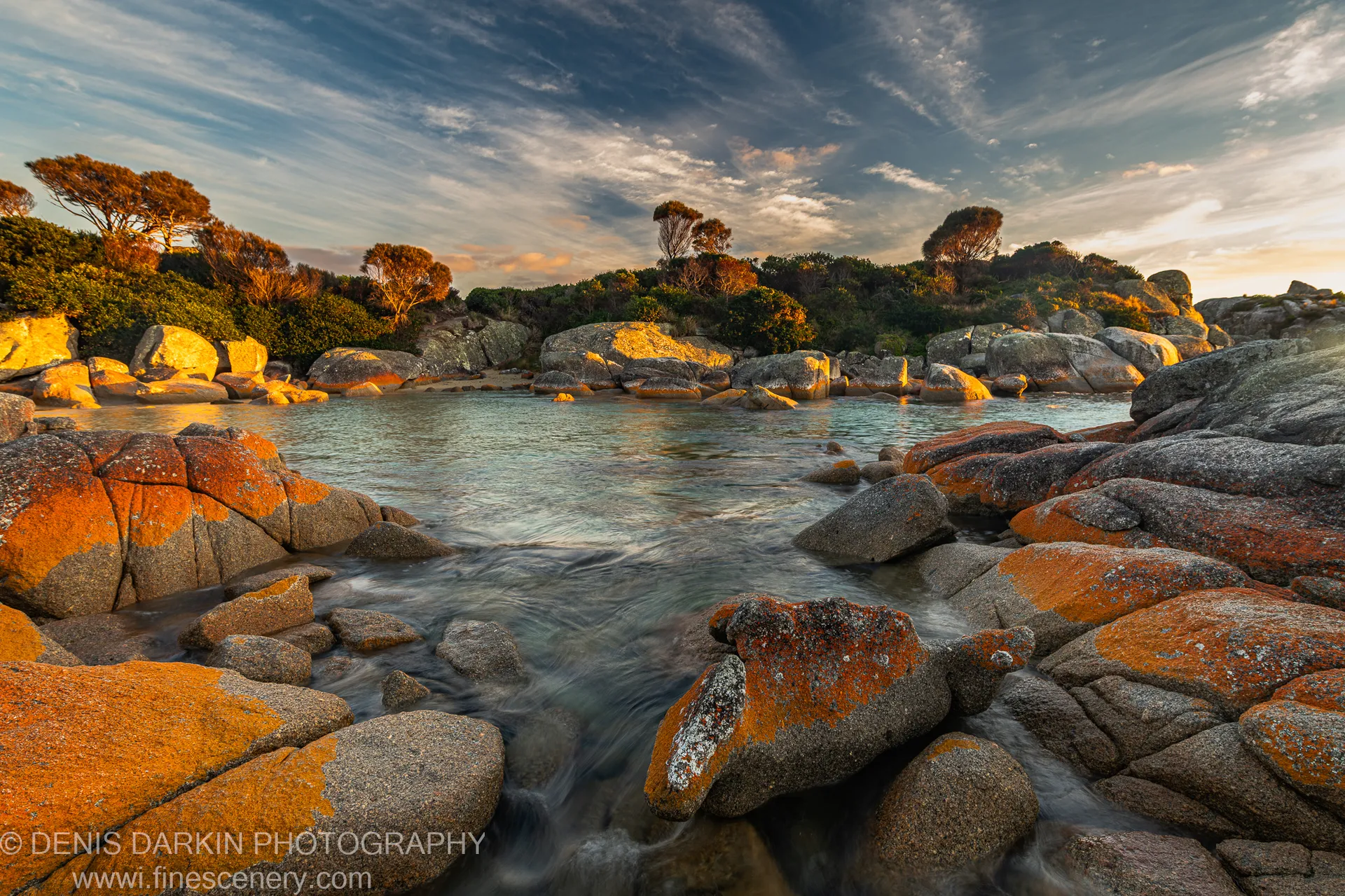 Shallow stream flows through a rocky channel lined with moss-covered boulders at Bay of Fires on a clear autumn morning.