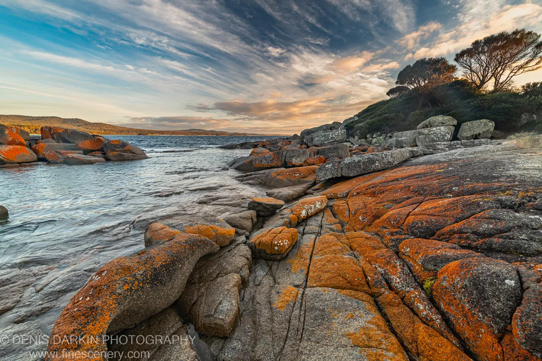 Wide-angle view of orange lichen-encrusted granite boulders at a tidal inlet, with cirrus clouds above and distant vegetated hills on the horizon.
