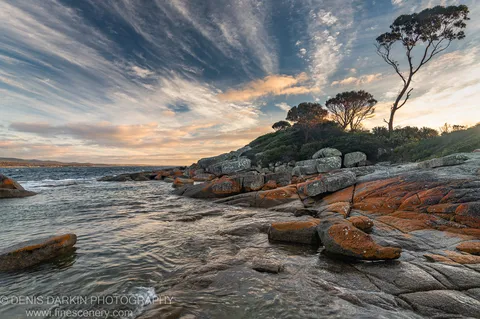 Granite boulders speckled with orange lichen line a shallow bay, with cool blue water and a distant warm horizon in late autumn.