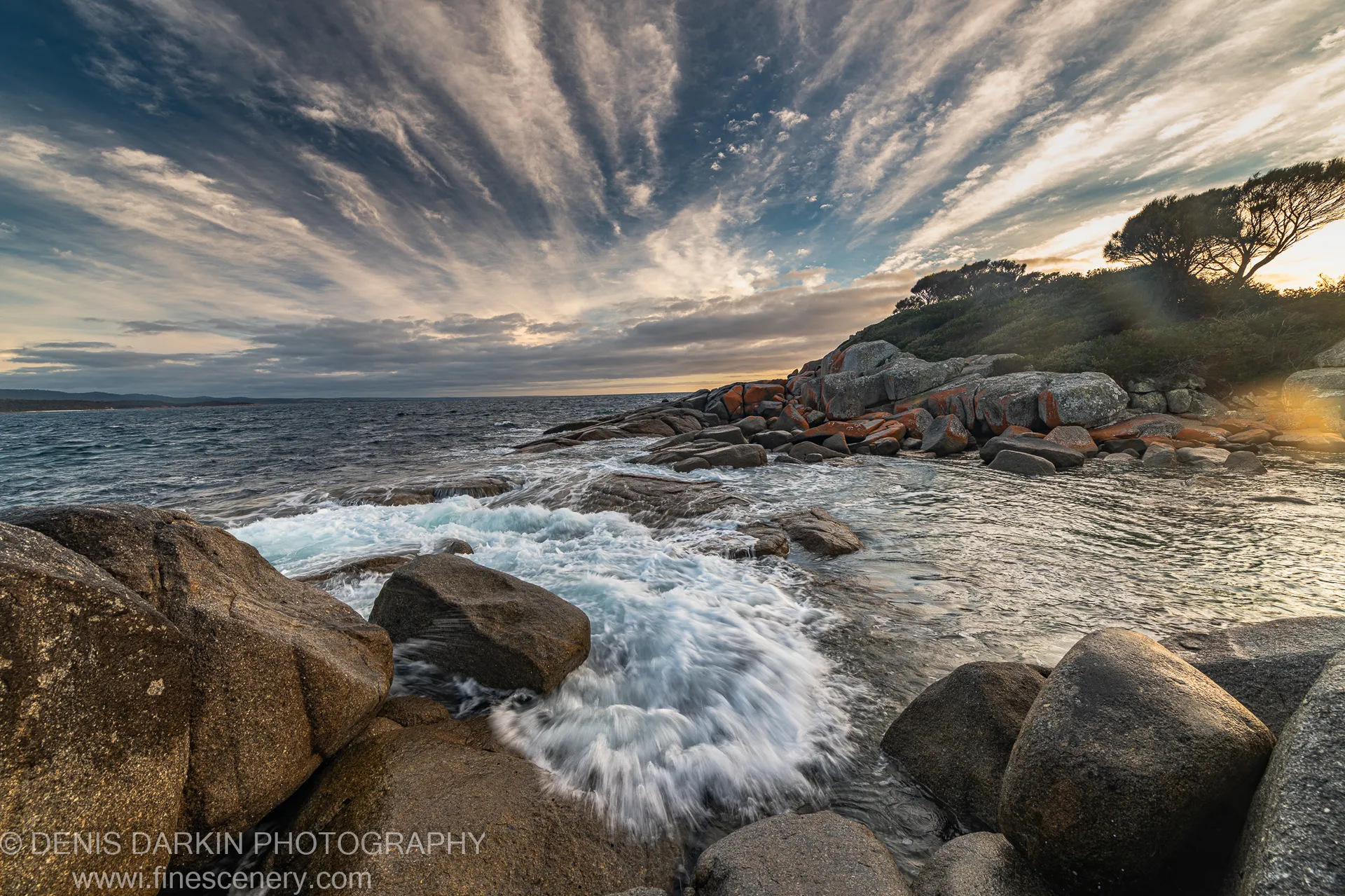 Radiating cirrus cloud bands stretch over a headland at Bay of Fires, granite boulders in the foreground lit by low morning sun.