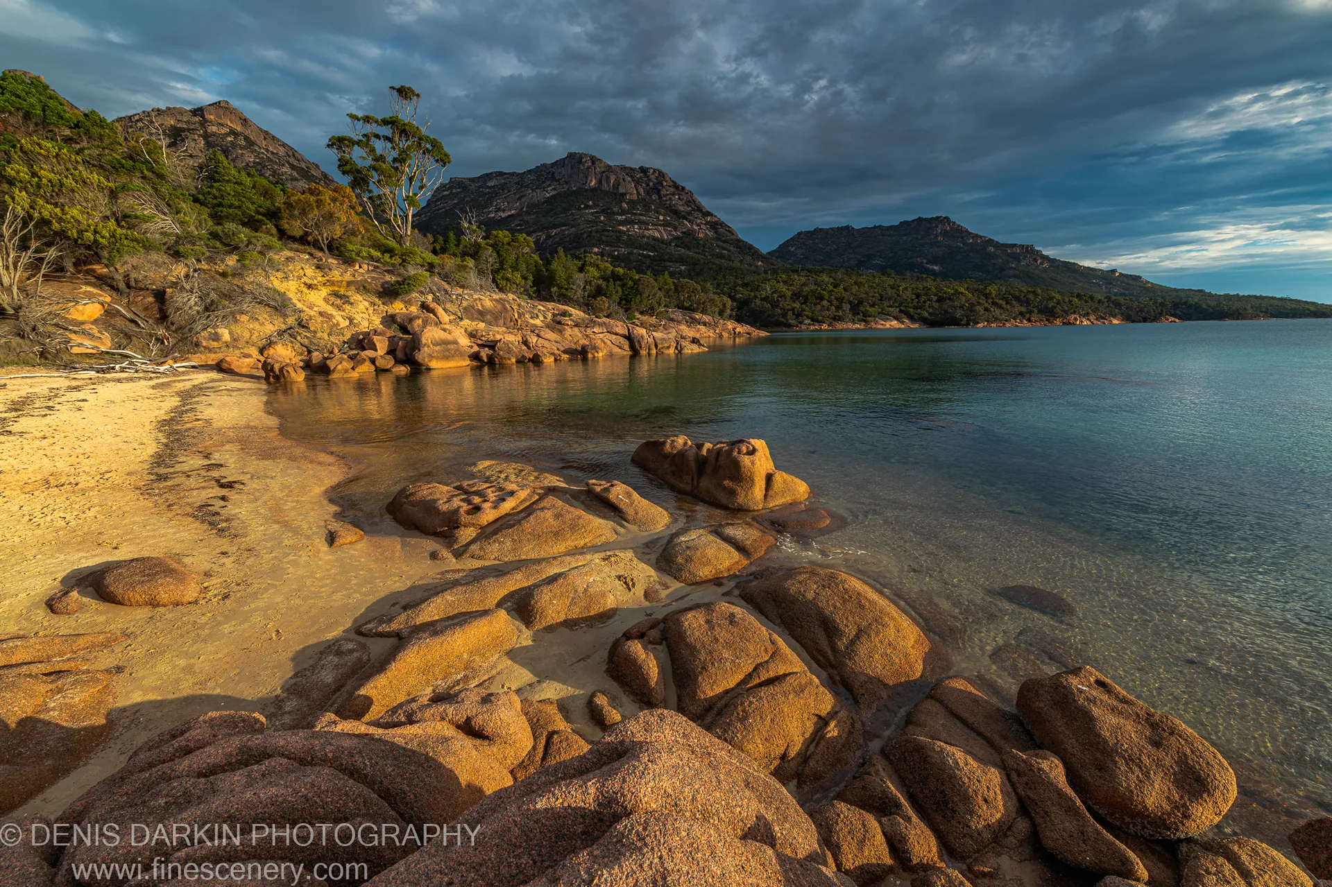 Sandy shoreline curving through scattered granite boulders toward the rugged peaks above Coles Bay, Tasmania, at golden hour in April 2022.