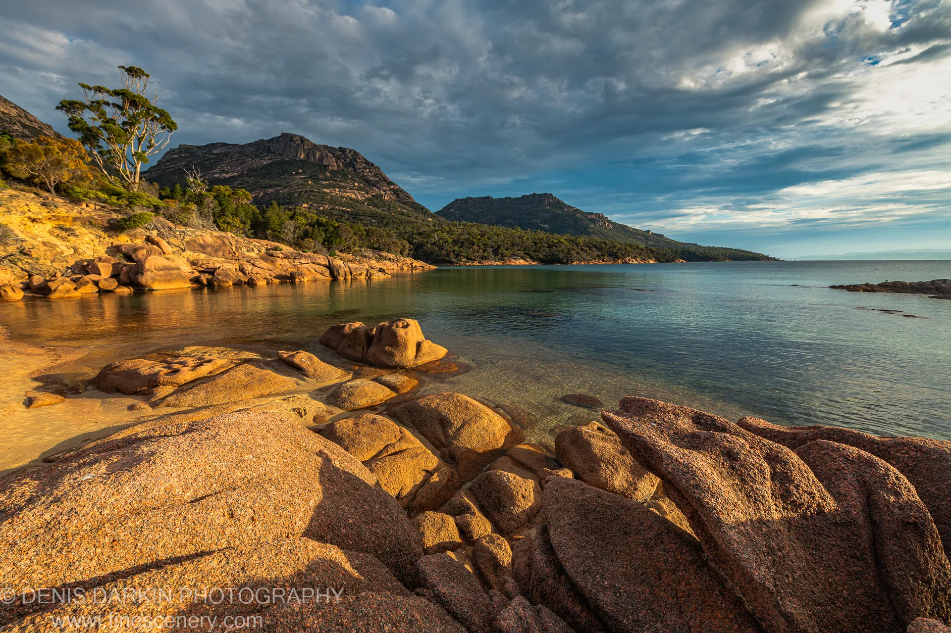 Massive granite boulders in warm light beside a glassy inlet, with cloud-streaked sky over scrub-covered hills at Coles Bay, April 2022.