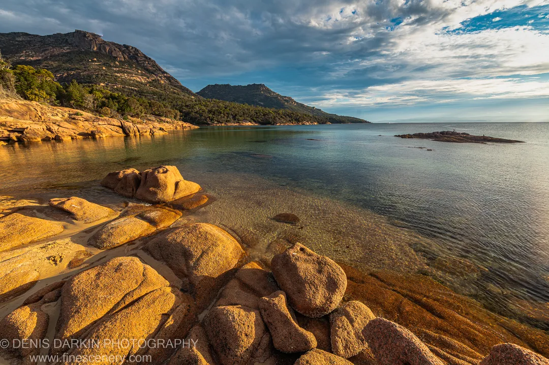 Sun-lit granite boulders above a clear shallow bay at Coles Bay, with steep tree-covered hills under textured clouds, April 2022.