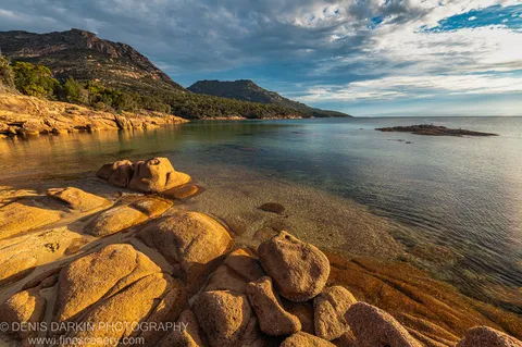 Sun-lit granite boulders above a clear shallow bay at Coles Bay, with steep tree-covered hills under textured clouds, April 2022.