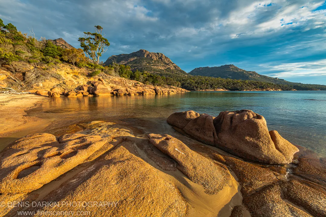 Massive rounded granite boulders in late-afternoon light at Coles Bay, with rugged mountain slopes and cloud banks above the still water beyond.