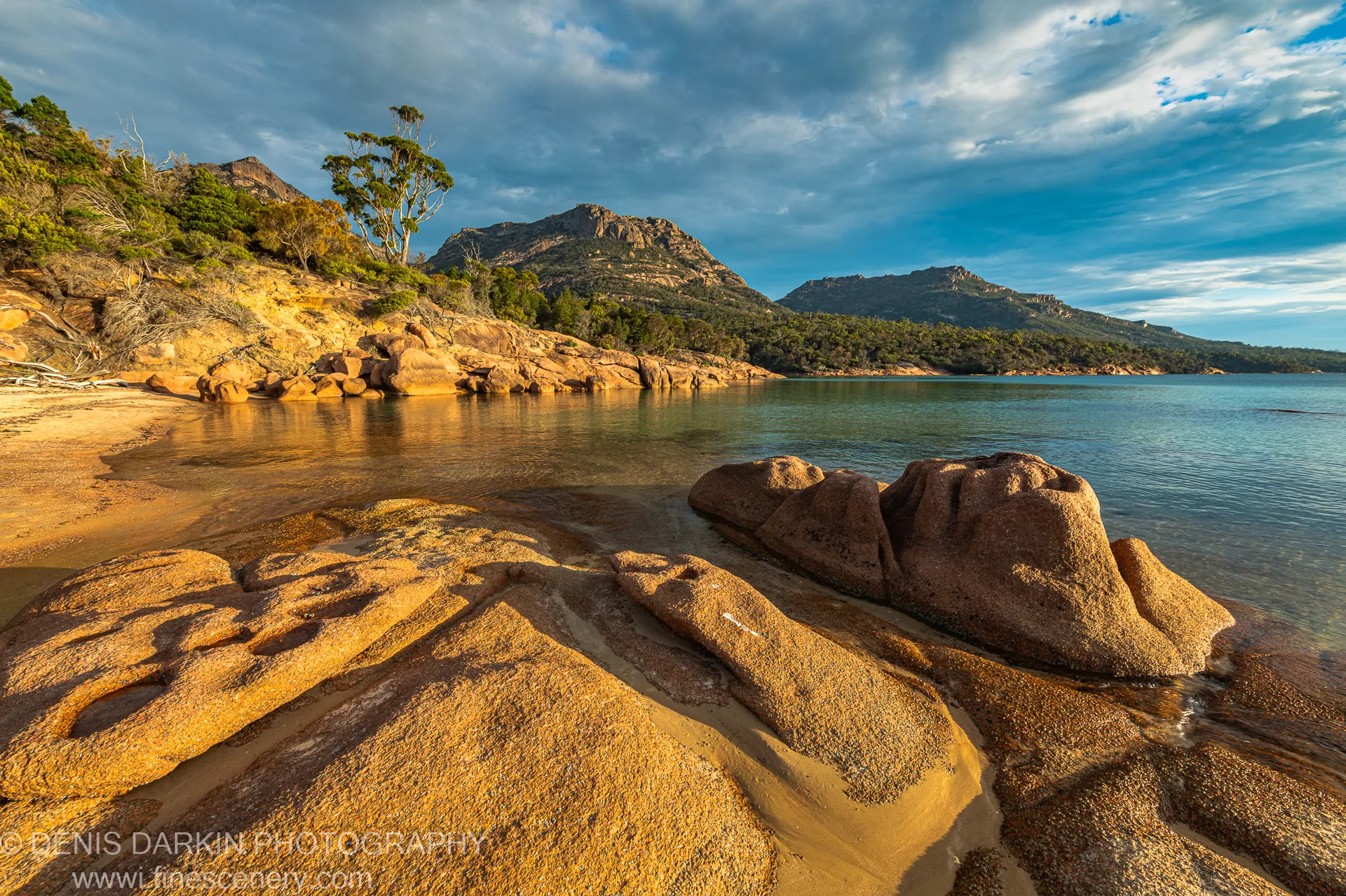Massive rounded granite boulders in late-afternoon light at Coles Bay, with rugged mountain slopes and cloud banks above the still water beyond.