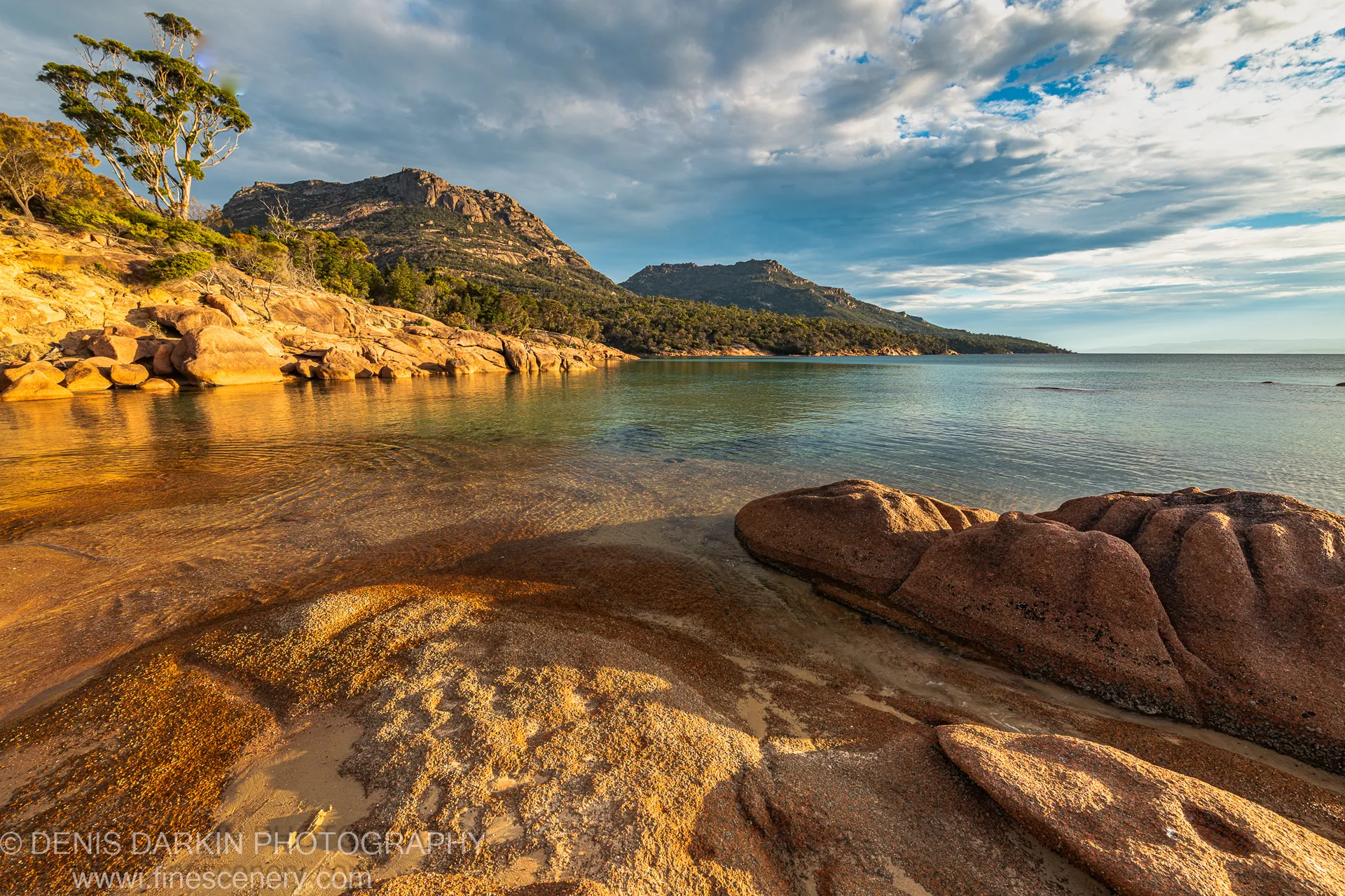 A windswept tree and a rock formation stand on the Coles Bay foreshore, with a distant headland visible below billowing clouds, April 2022.