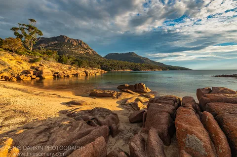 Warm afternoon light on rough granite boulders at the edge of a sheltered cove in Coles Bay, with textured clouds over the rugged hillside.