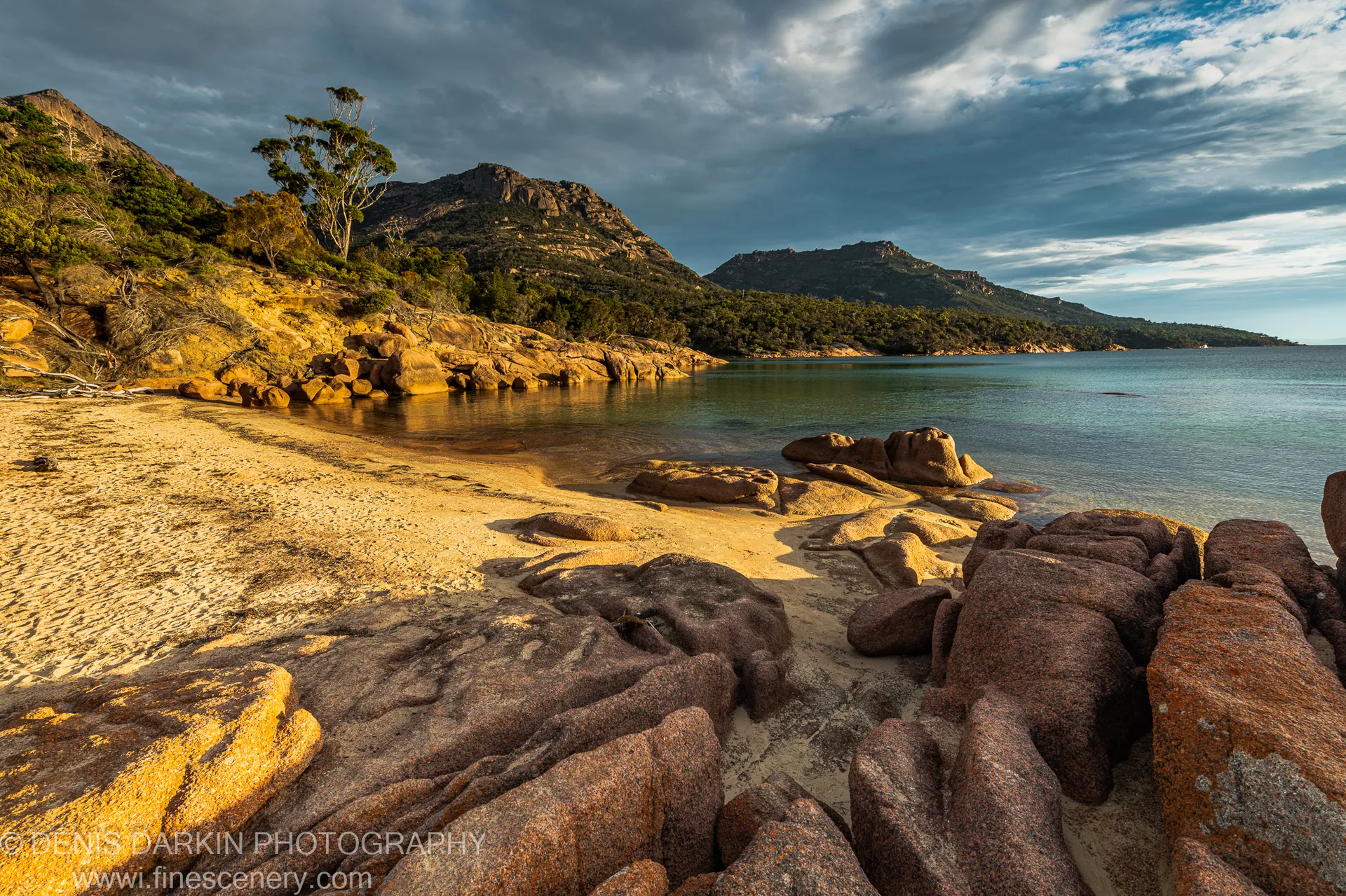 Sunlit granite boulders and sandy beach at Coles Bay, Tasmania, beneath a heavy sky of slate-grey cloud, with dark-green coastal vegetation on the distant slopes.