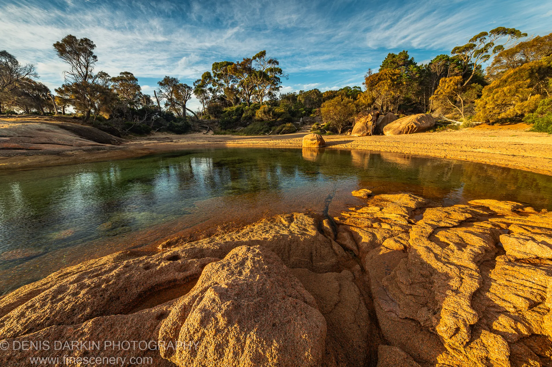 Sun-lit granite boulders at the edge of a calm inlet at Coles Bay, Tasmania, where clear water reflects the silhouettes of coastal trees.