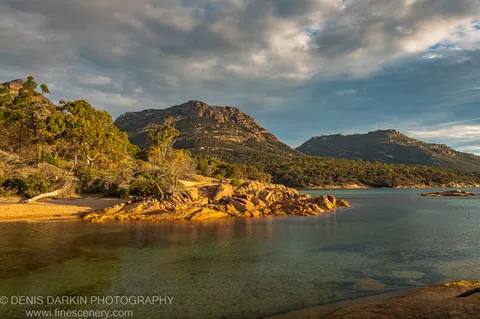Afternoon sun on granite outcrops and coastal foliage at Coles Bay, with heavy cloud and blue sky breaks above the rugged mountain shoreline.