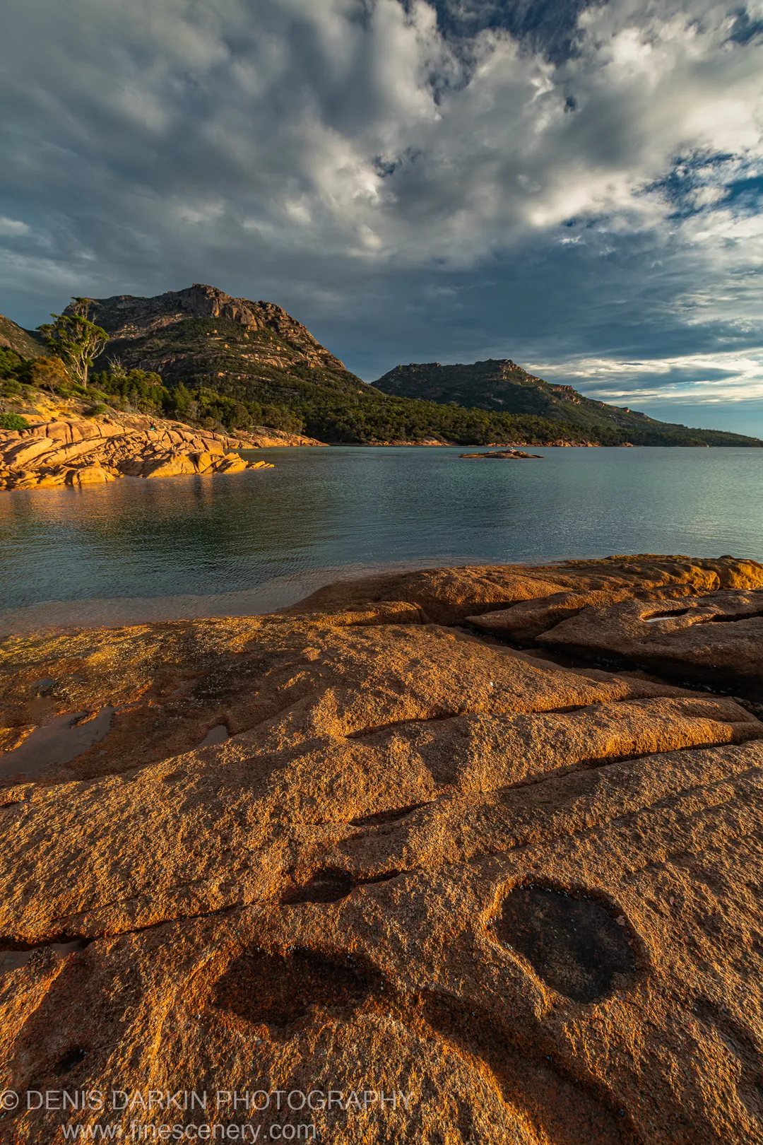 Sunlit granite rocks stretching across the foreground of a calm Tasmanian bay, with steep forested headlands and a dark heavy sky at golden hour.
