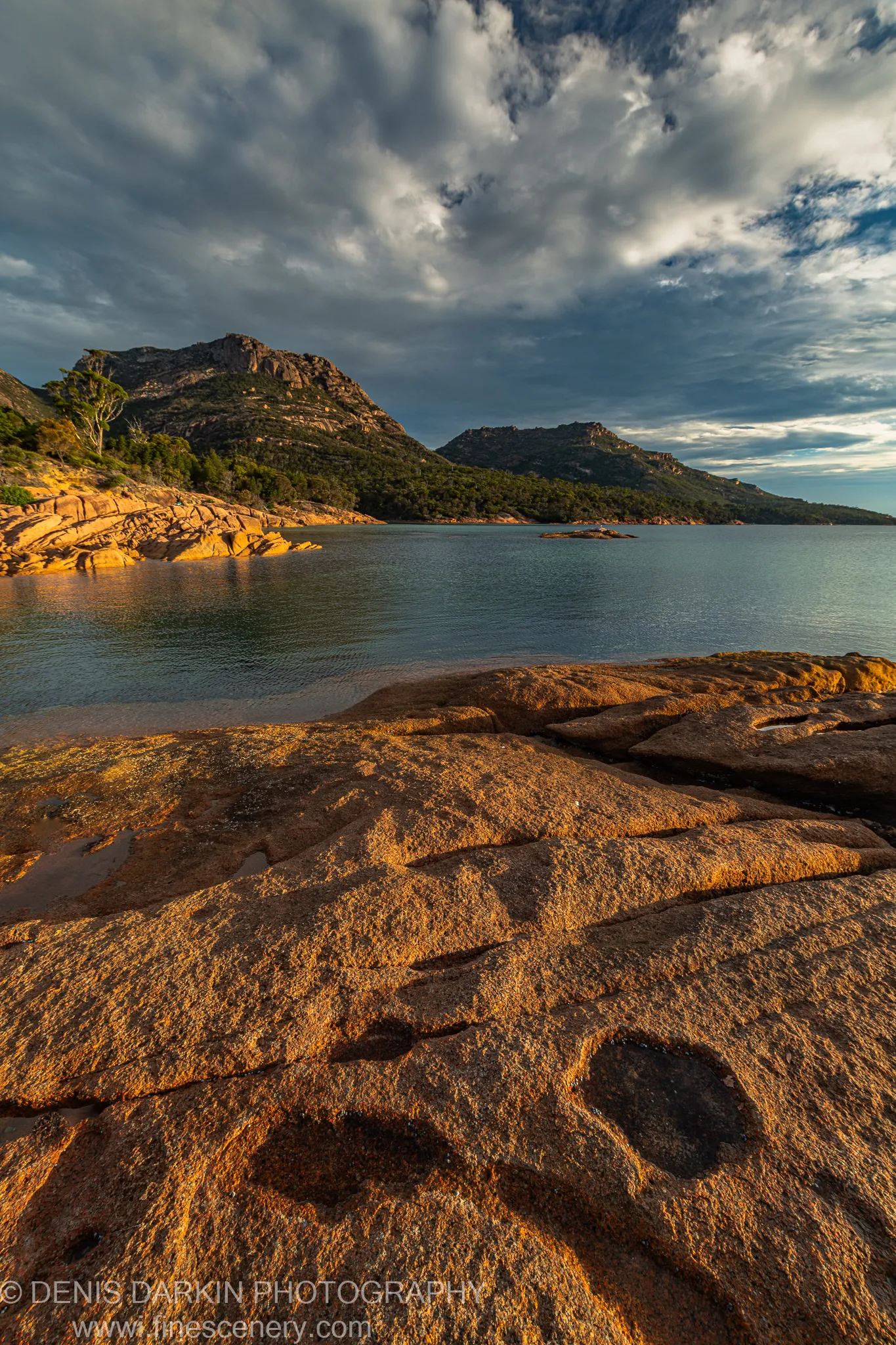 Sunlit granite rocks stretching across the foreground of a calm Tasmanian bay, with steep forested headlands and a dark heavy sky at golden hour.