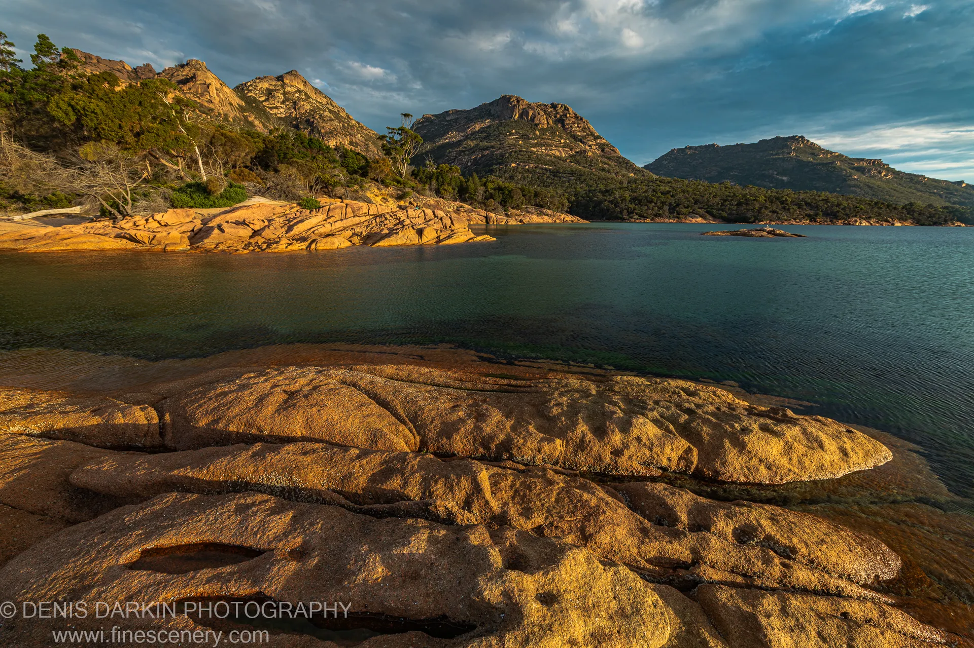 Wet granite rocks at the edge of a blue-green inlet at Coles Bay, with rugged mountain peaks and a cloud-streaked sky in the distance.