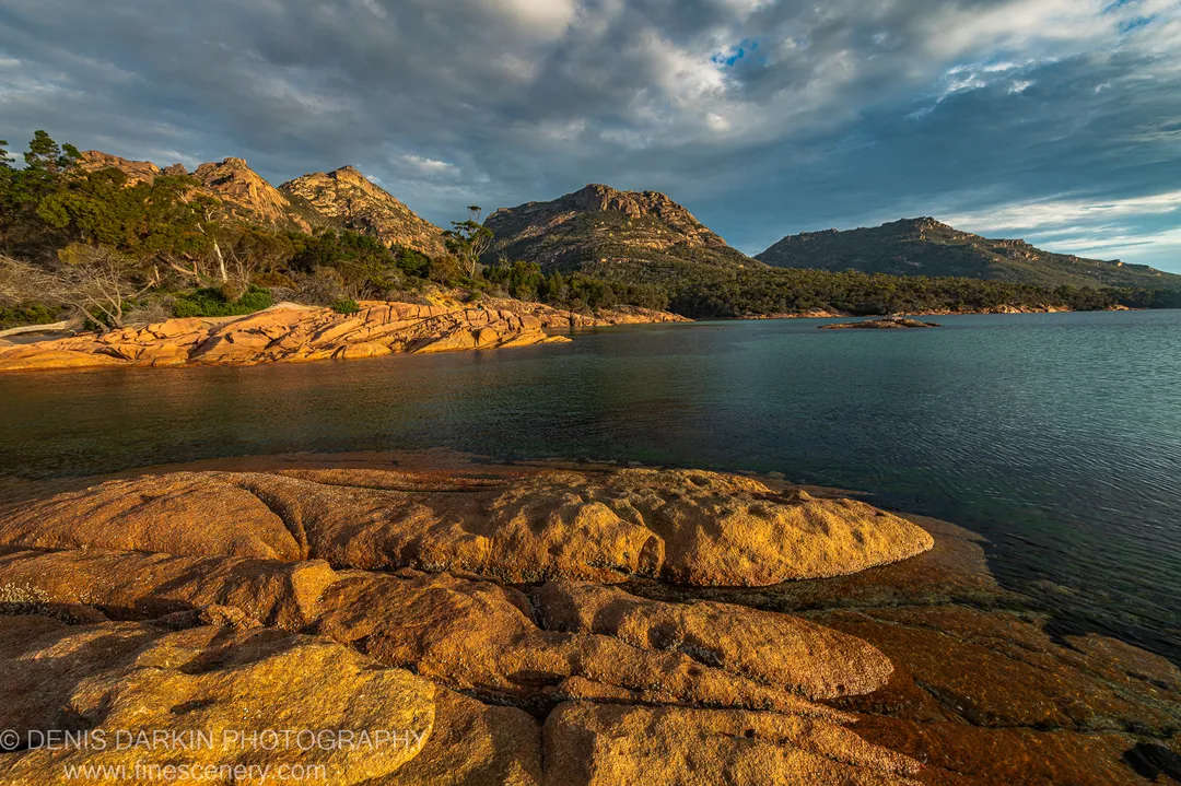 Lichen-covered weathered granite on the Coles Bay foreshore, with calm water leading to rugged mountain peaks beneath a sky of long streaking cloud.