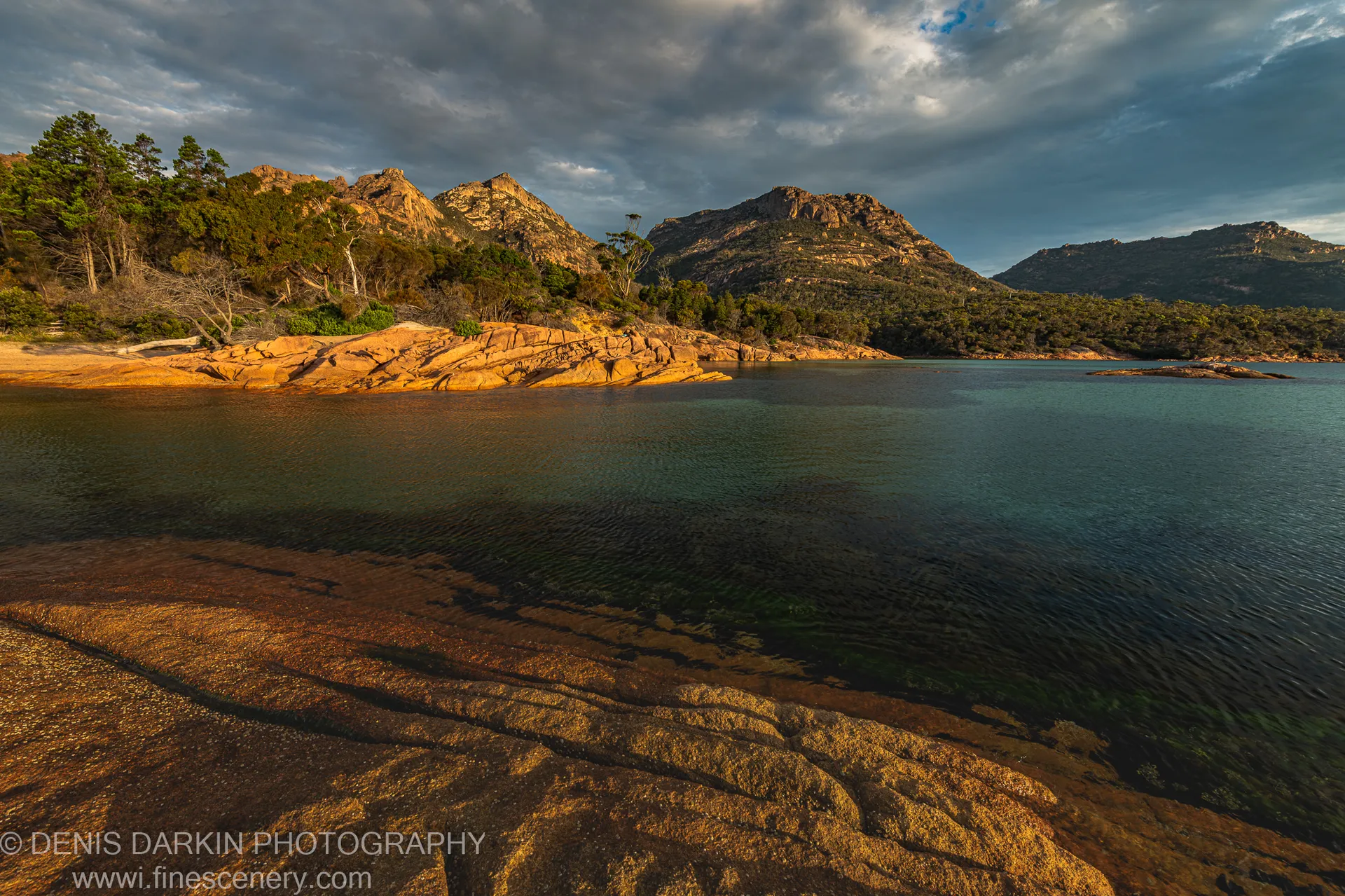 Stratified rock formations rising from shallow water at Coles Bay, Tasmania, the warm amber foreground contrasting with dark mountain slopes and a heavy layered sky.