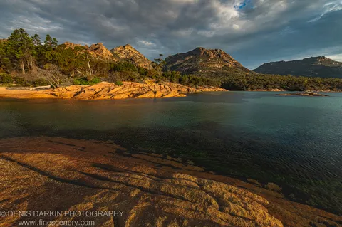 Stratified rock formations rising from shallow water at Coles Bay, Tasmania, the warm amber foreground contrasting with dark mountain slopes and a heavy layered sky.