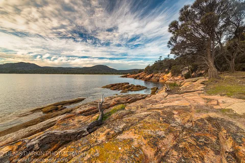 Lichen-flecked granite boulders at Coles Bay, Tasmania, catch warm late-afternoon light, with calm water and forested hills beyond.