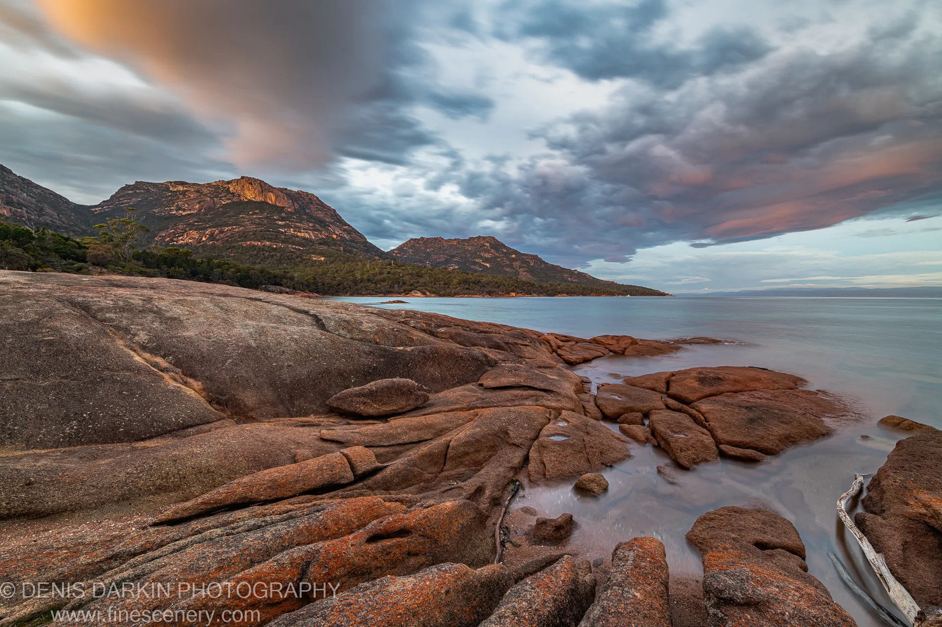 Smooth granite boulders form a foreshore platform at Coles Bay, Tasmania, with long-exposure water and distant mountains visible on a late-autumn morning.