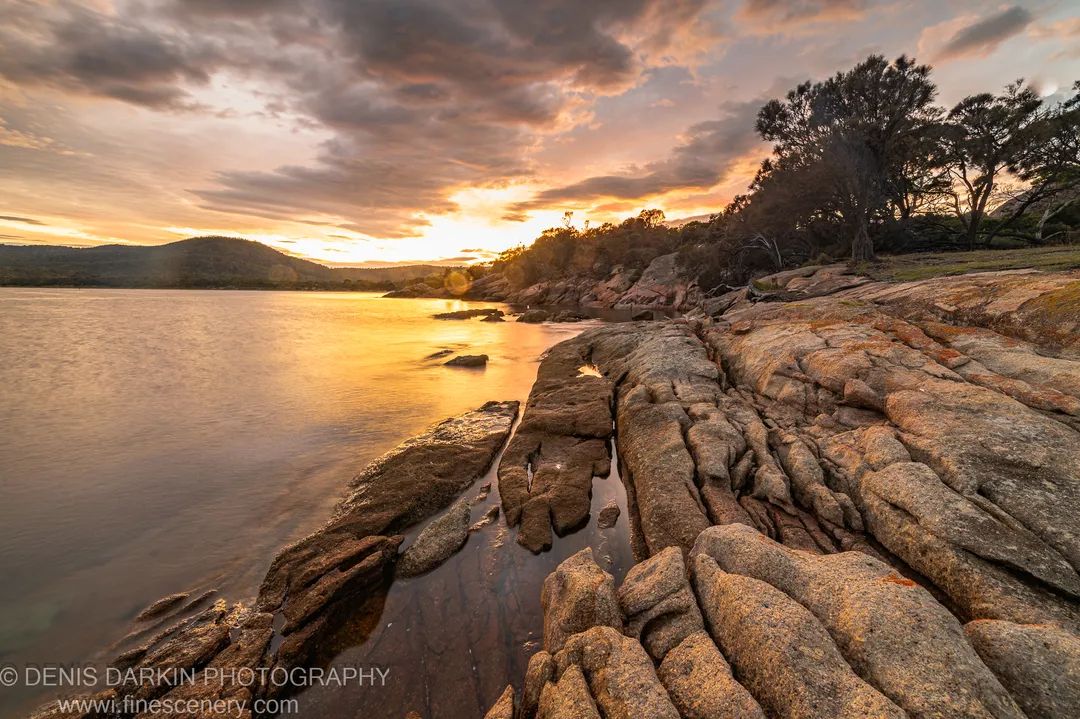 Smooth layered rock formations at Coles Bay, Tasmania, extend toward still long-exposure water below a textured cloud bank on an autumn morning.