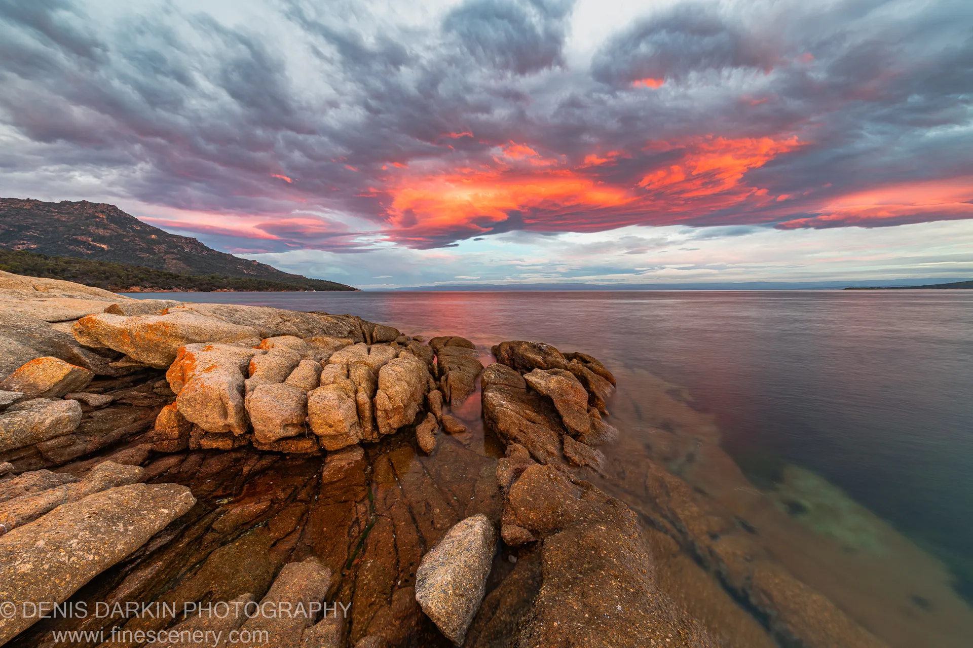Orange-lichened granite boulders line the shore at Coles Bay, Tasmania, beneath a sky of billowing cloud lit orange in early morning.