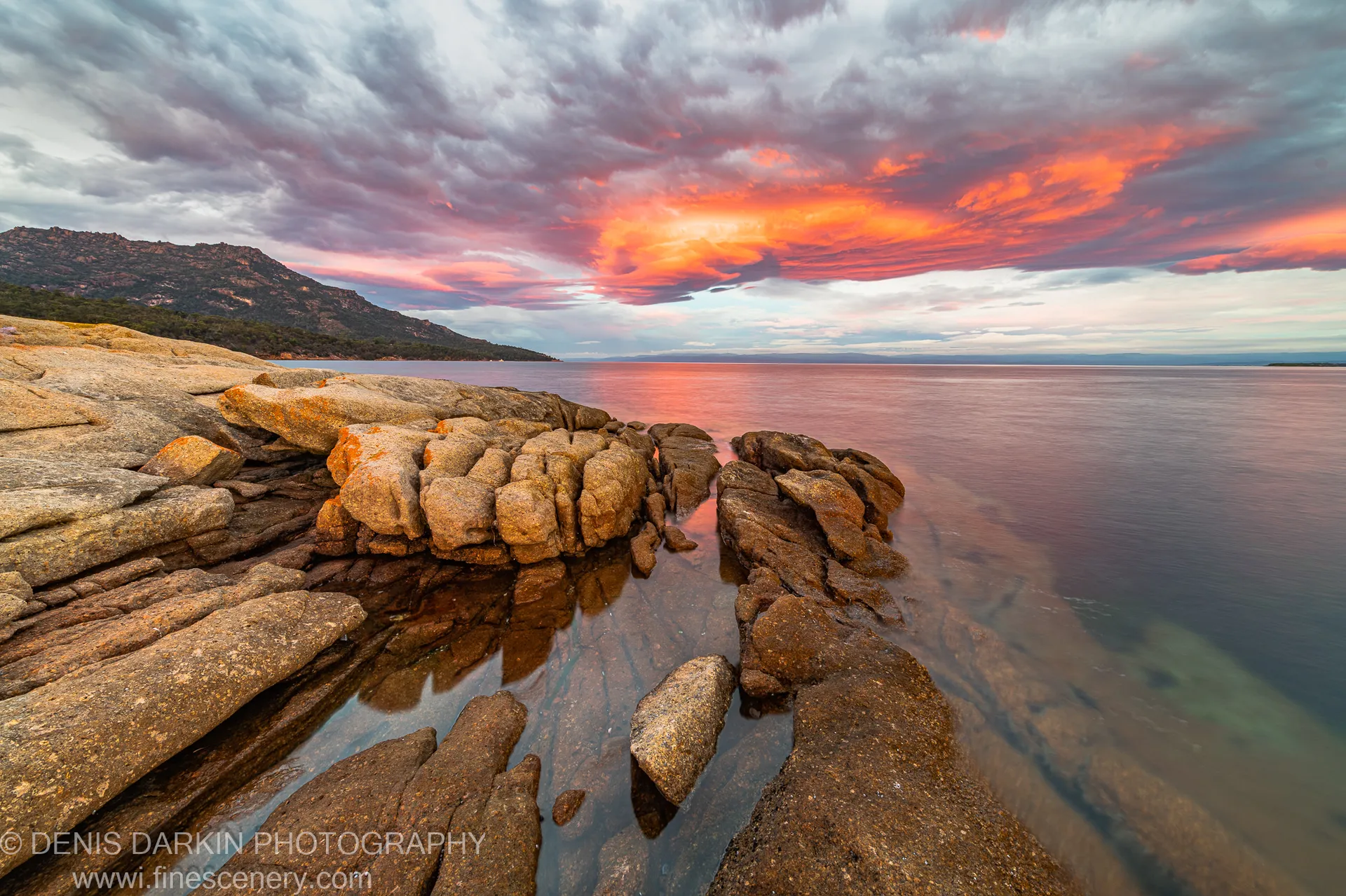 Rugged granite boulders at the shore of Coles Bay, Tasmania, with ocean water smoothed by long exposure beneath a pink and orange morning sky.