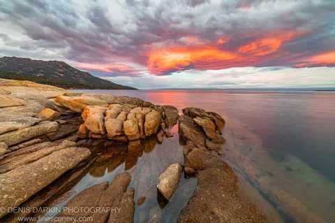 Rugged granite boulders at the shore of Coles Bay, Tasmania, with ocean water smoothed by long exposure beneath a pink and orange morning sky.