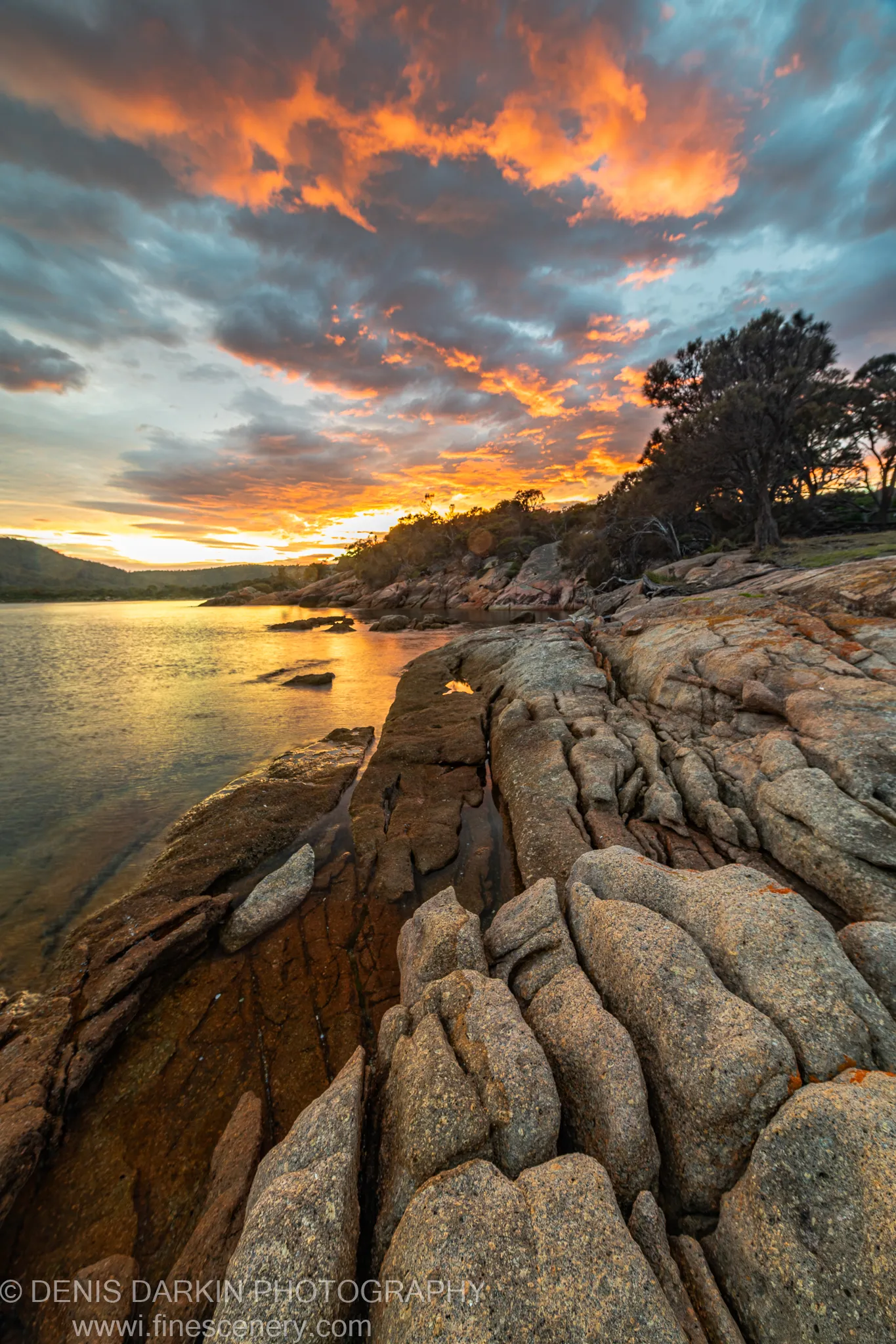 Layered granite formations at the water's edge, Coles Bay, Tasmania, with clouds lit orange and gold above the calm bay in morning light.