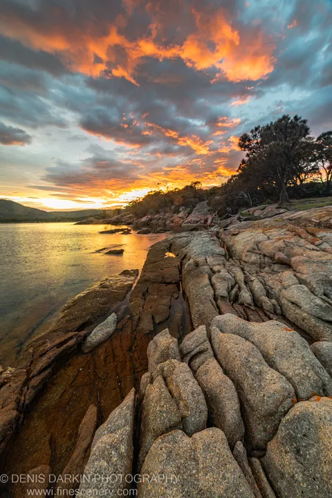 Layered granite formations at the water's edge, Coles Bay, Tasmania, with clouds lit orange and gold above the calm bay in morning light.