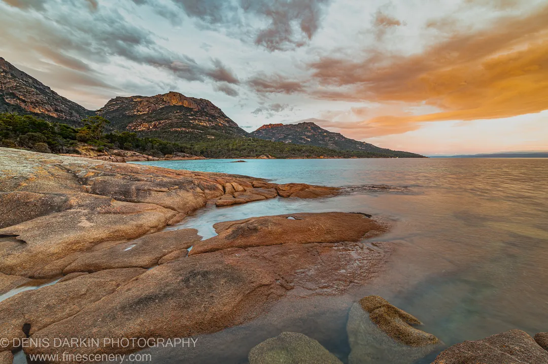 Smooth granite slabs in the foreground at Coles Bay, Tasmania, with the glassy bay and twin tree-covered peaks of the Hazards beyond.
