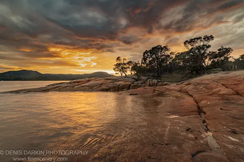 Smooth granite boulders at the water's edge at Coles Bay, Tasmania, the bay reflecting orange light from the morning sky above dark cloud.