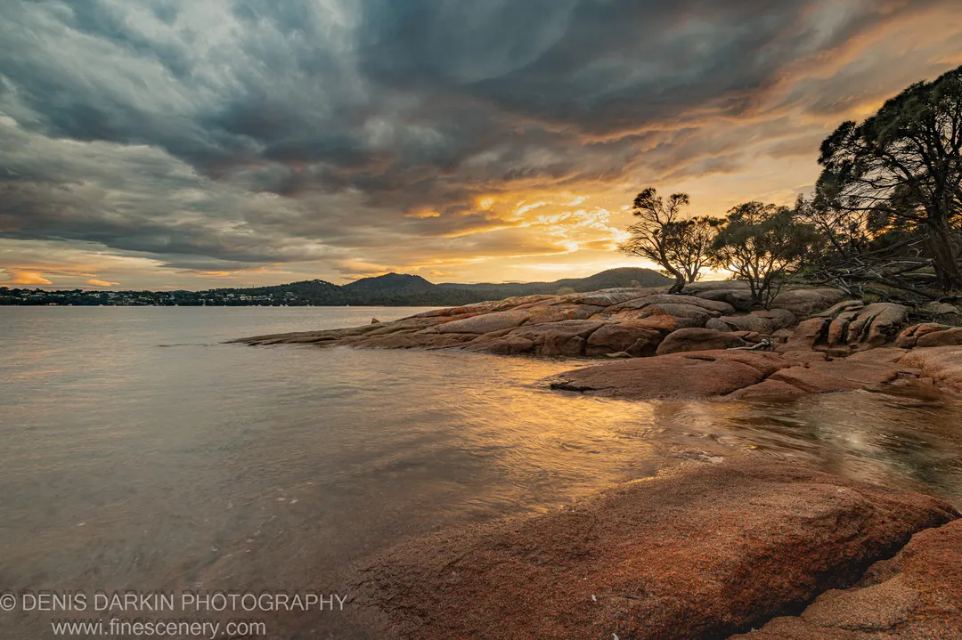 Textured granite boulders on the shore at Coles Bay, Tasmania, beneath rolling storm cloud, a band of morning light breaking at the horizon.
