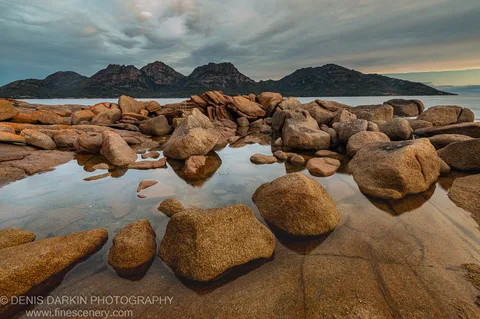 Large granite boulders covered in orange lichen rest in clear tidal pools below a mountain silhouette at Coles Bay, East Coast Tasmania.