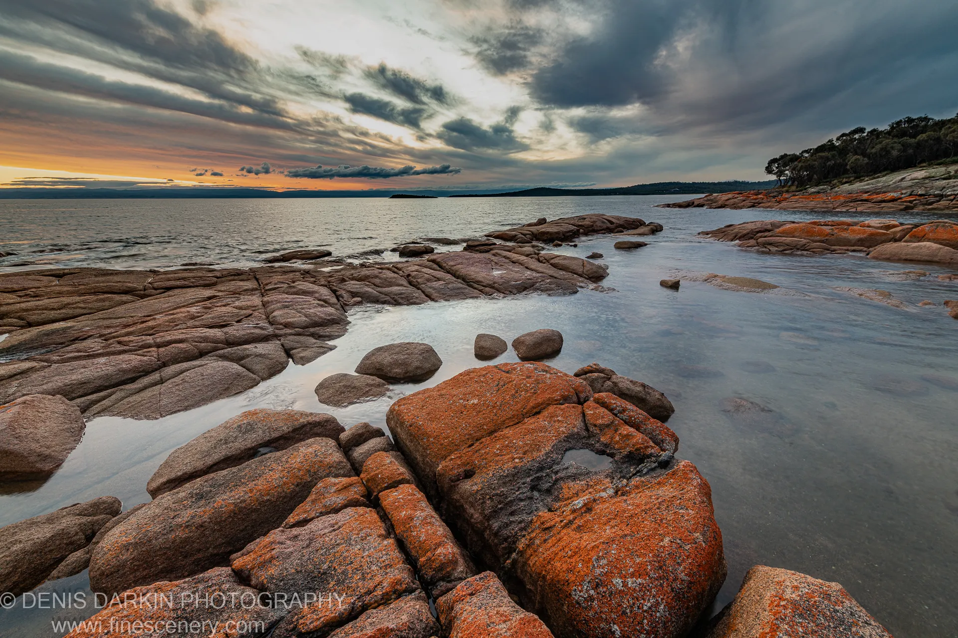 Orange lichen on granite boulders at Coles Bay, calm pools in the foreground and a distant headland silhouetted against amber light at sunset.