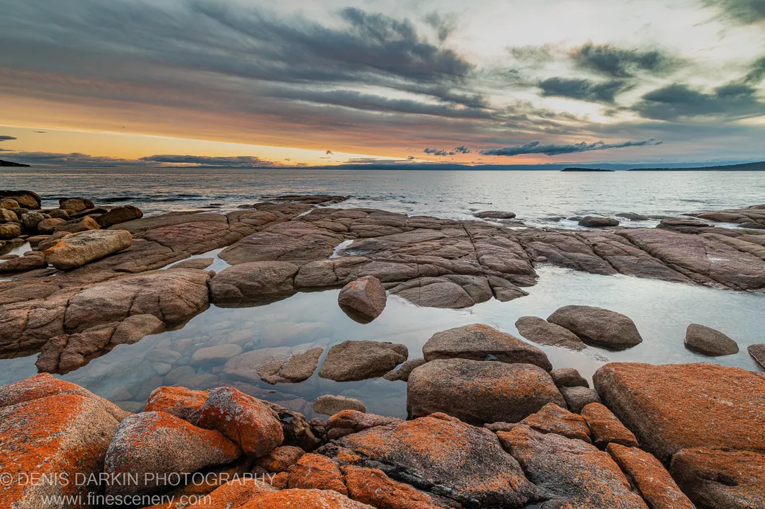 Orange lichen on granite boulders at Coles Bay, storm clouds above and an amber glow at the horizon, distant landmasses in silhouette.