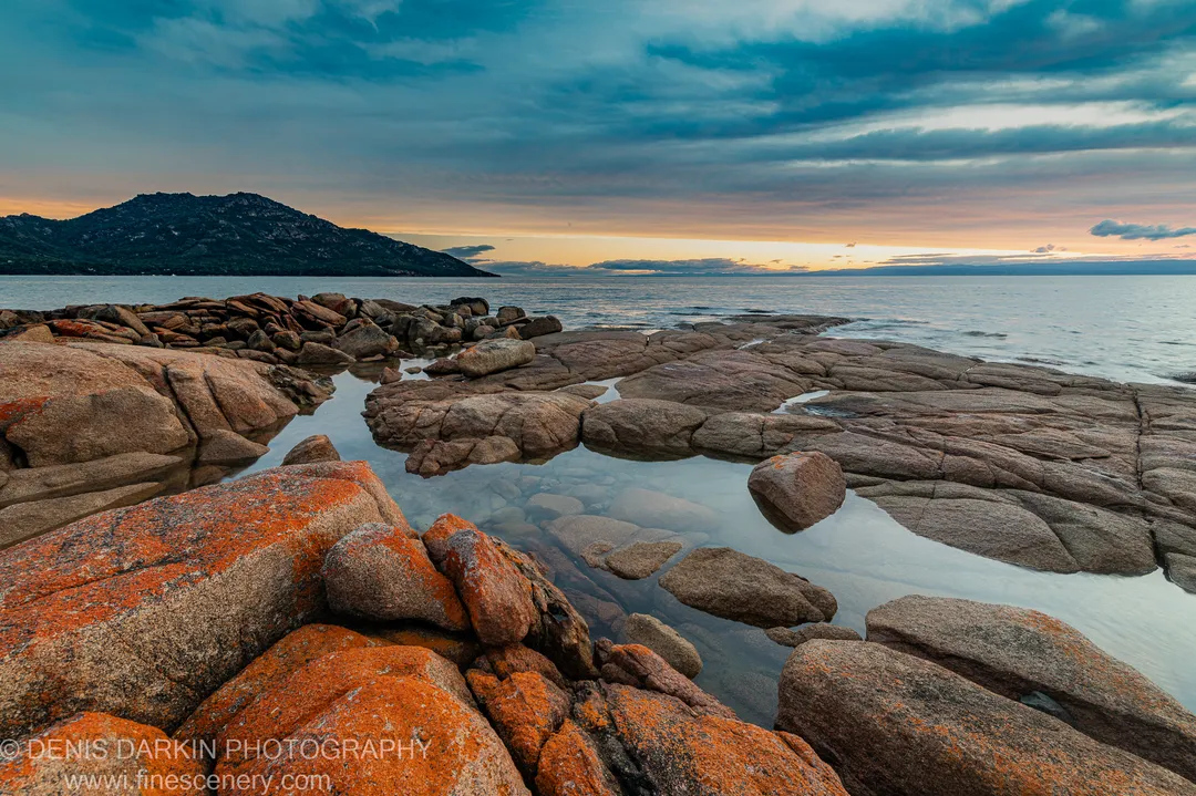 Granite boulders covered in orange lichen edge calm tide pools at Coles Bay, with smooth ocean stretching toward a distant headland under a gradient sky.