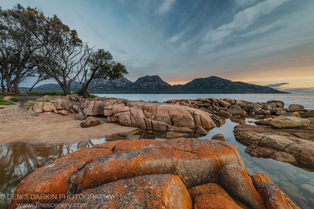 Large granite boulders covered in orange lichen at Coles Bay, a sandy beach and dark craggy mountains rising against a streaked sky.