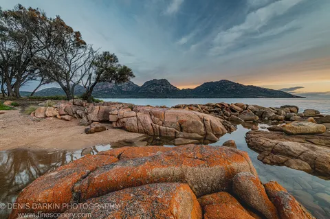 Large granite boulders covered in orange lichen at Coles Bay, a sandy beach and dark craggy mountains rising against a streaked sky.