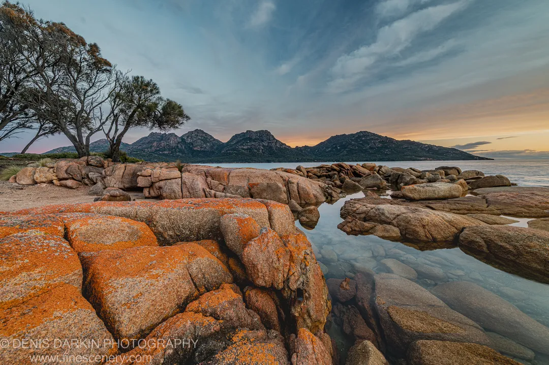 A clear rock pool reflects orange lichen-covered granite boulders at Coles Bay, with a dark headland and soft sunset glow at the horizon.