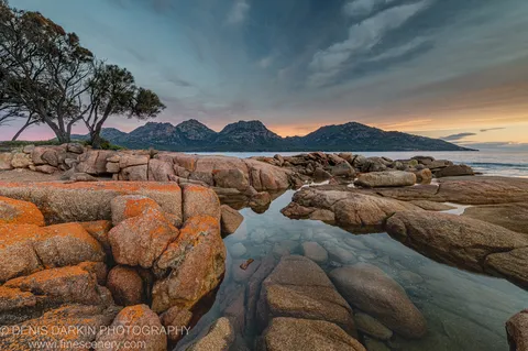 Orange lichen on smooth granite at Coles Bay, with tidal pools in the foreground and the Hazards rising beyond the still water at golden hour.