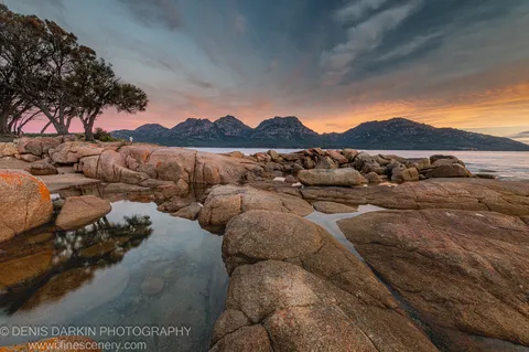 Dark coastal tree trunks and the warm tones of the evening sky reflected in clear tidal pools among granite boulders at Coles Bay.
