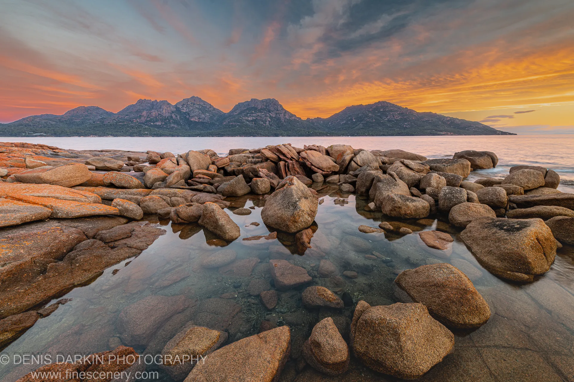 Lichen-textured granite boulders in warm sunset light at Coles Bay, with jagged mountain peaks silhouetted against an orange and violet streaked sky.