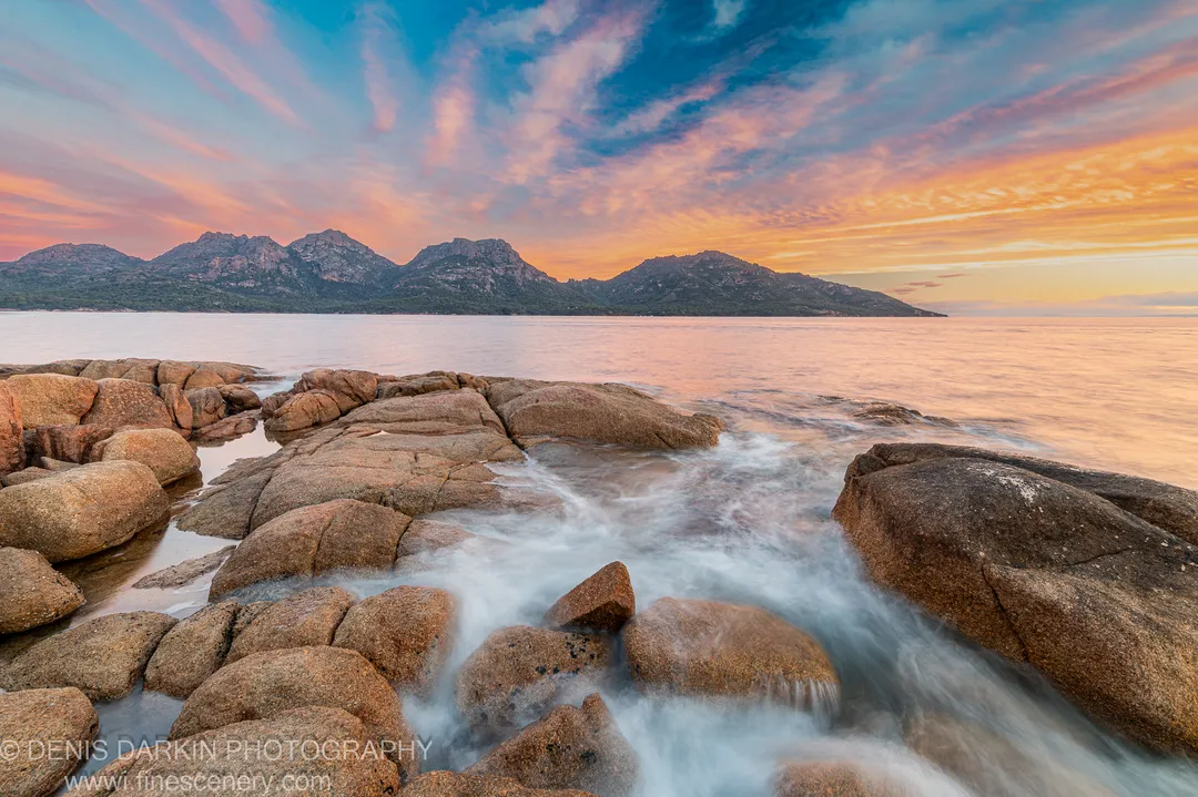 Large rounded granite boulders with water flowing between them at Coles Bay, the Hazards mountains rising under a pink and blue streaked sky at sunset.