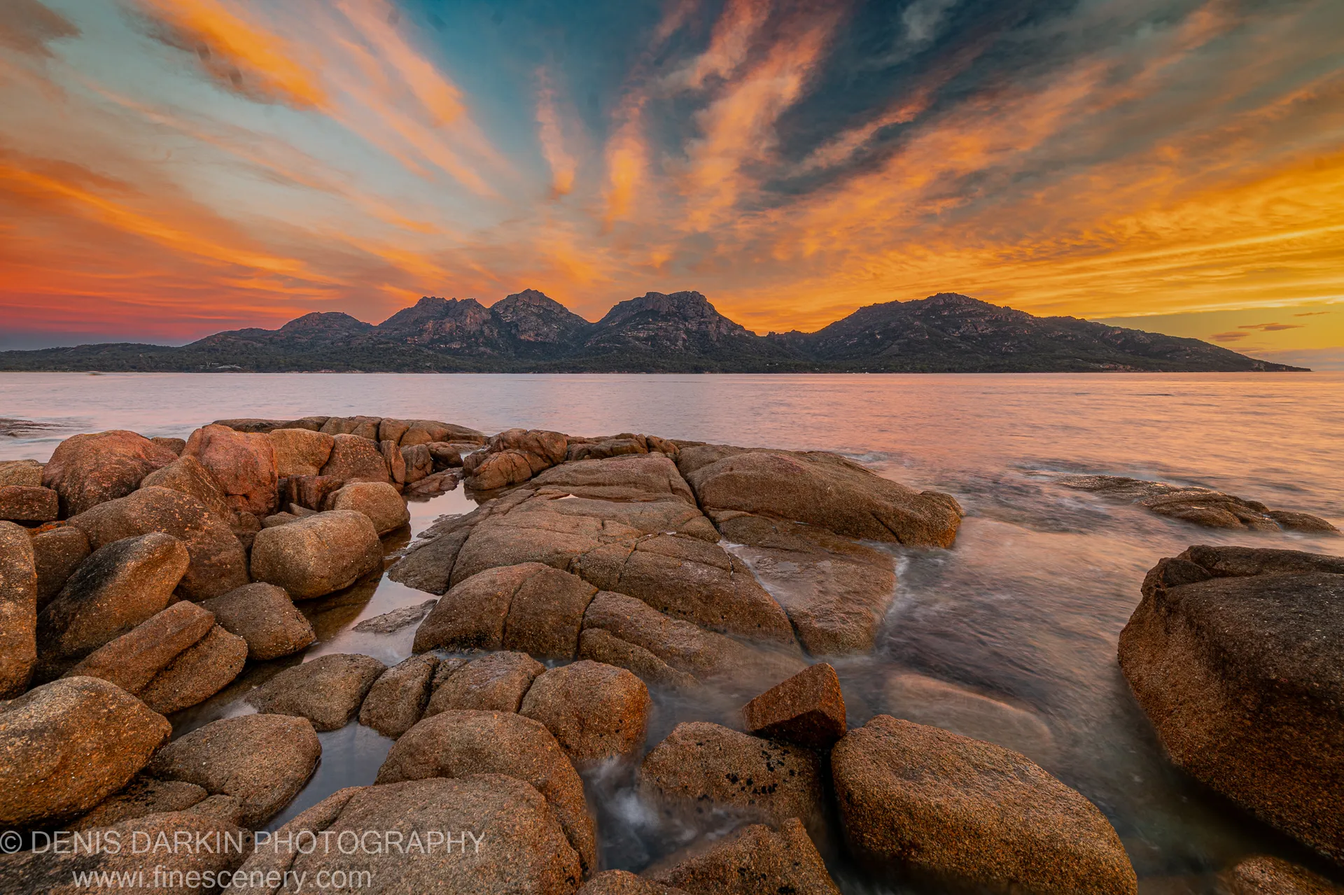 Rounded granite boulders with motion-blurred water in the foreground at Coles Bay, a dark mountain range silhouetted against a sunset sky of orange and blue.