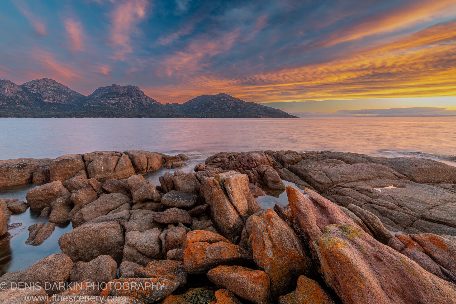 Lichen-streaked granite boulders in warm light at Coles Bay, with apricot and violet clouds over calm water and the Hazards in the distance.